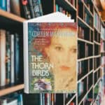 The cover features a woman with red hair flowing behind her. In the bottom corner there is an image of a house. Behind the cover are shelves of books.