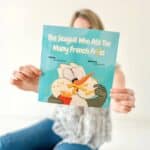 Author Ocean Atwood sitting on the ground with her book, The Seagull Who Ate Too Many French Fries covering her face. The cover shows a cartoon seagull shoving french fries into its beak.