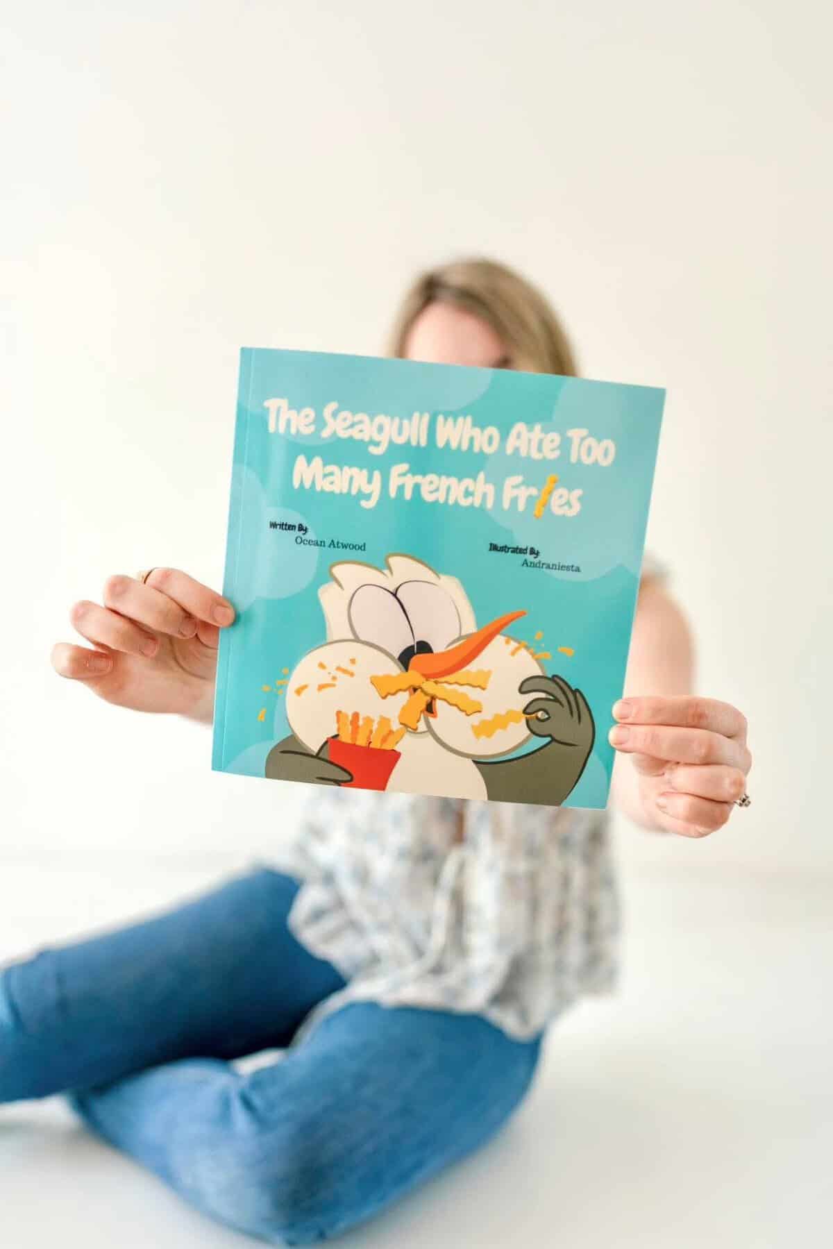 Author Ocean Atwood sitting on the ground with her book, The Seagull Who Ate Too Many French Fries covering her face. The cover shows a cartoon seagull shoving french fries into its beak.