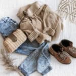 Women's fall clothing folded on a bed. The outfit includes, jeans, boots, a knitted sweater, and a small wicker handbag.