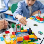 Two boys play with LEGO at a table. A boy in a blue shirt reaches his LEGO person towards the other boy's building.