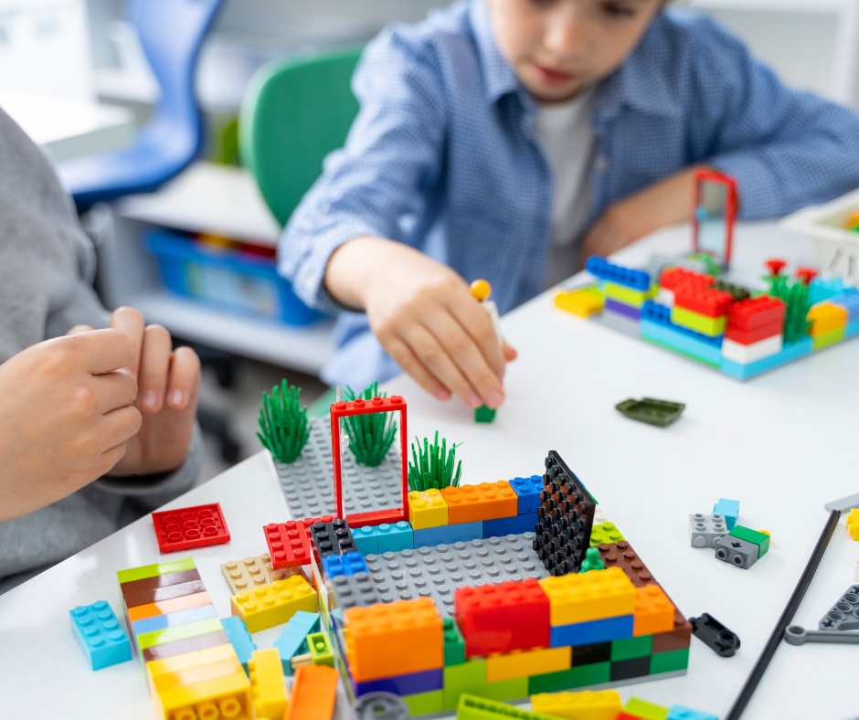Two boys play with LEGO at a table. A boy in a blue shirt reaches his LEGO person towards the other boy's building.