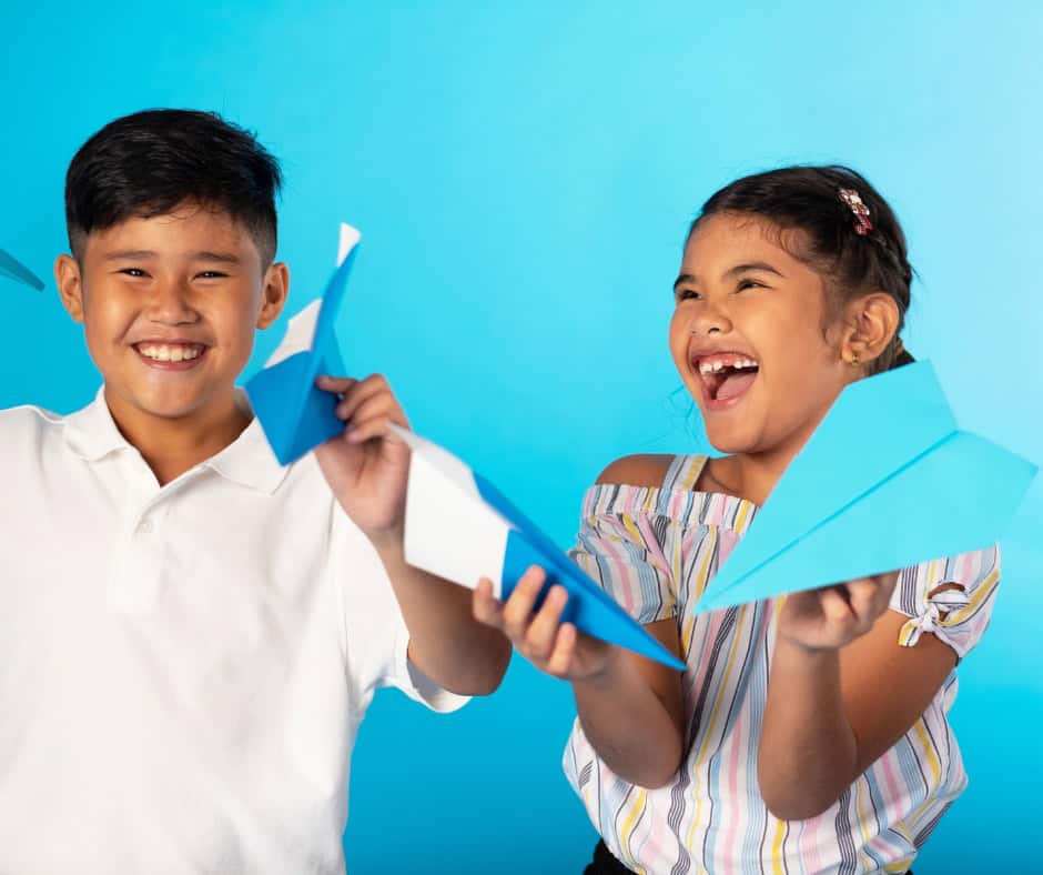 A boy and girl smiling and holding paper airplanes in their hands. The background is blue and so are the airplanes.