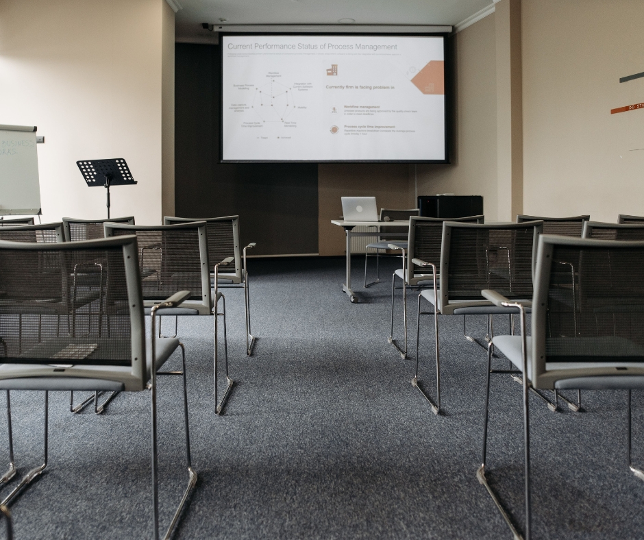 A room lined with metal chairs. At the back of the room is a projector screen with a presentation being shown.