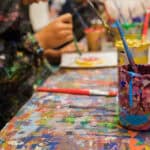 A photographed shot of a table covered in paint. There is a cup and paintbrushes in the foreground and children in the background.