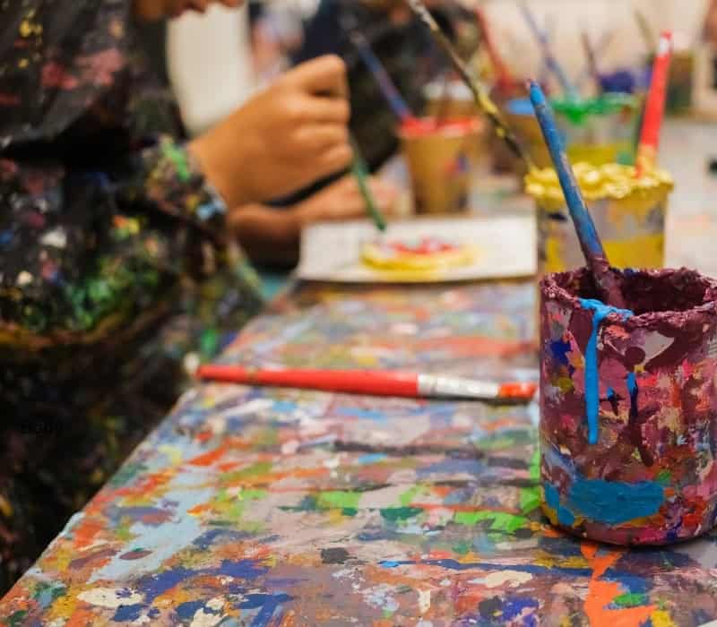 A photographed shot of a table covered in paint. There is a cup and paintbrushes in the foreground and children in the background.