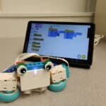 A little robot with eyes made out of LEGO Spike materials. Behind it is a tablet with the LEGO Spike app open and coding blocks visible.