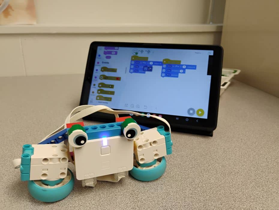 A little robot with eyes made out of LEGO Spike materials. Behind it is a tablet with the LEGO Spike app open and coding blocks visible.