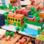 A colourful city made from cardboard, tissue paper, and other craft materials. In the background children are adding things to the city.