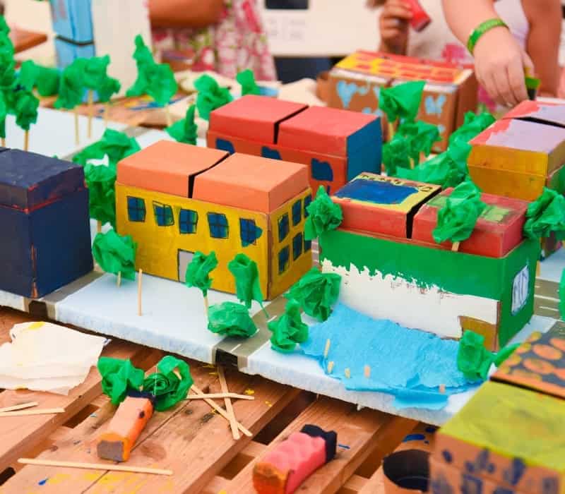 A colourful city made from cardboard, tissue paper, and other craft materials. In the background children are adding things to the city.