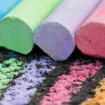 Sticks of chalk in blue, green, purple, pink, and orange. In front of them are lines drawn on concrete.