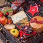 A wicker basket full of foods like homemade cookies, cheese, apples, and crackers. They are all wrapped in Christmas ribbon.