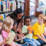 A group of children with different ethnicities. They each have a book in their hands and are listening to an African Canadian woman who speaks to them.