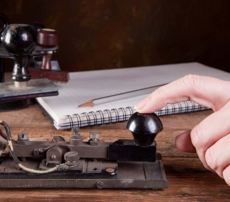 A hand using an old fashioned morse code machine to send a message. Beside it is a notebook and pen for writing down responses.