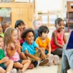 A group of young children with different ethnicities sitting on the floor and listening to a story. The woman reading the story is out of shot.
