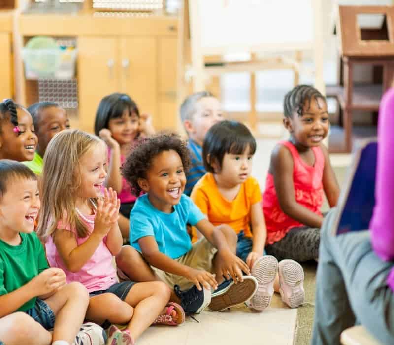 A group of young children with different ethnicities sitting on the floor and listening to a story. The woman reading the story is out of shot.