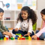 Three preschool-aged children playing with LEGO. There is a boy with short dark hair on the left, a girl with long curly hair in the middle, and a girl with straight hair in a ponytail on the right.