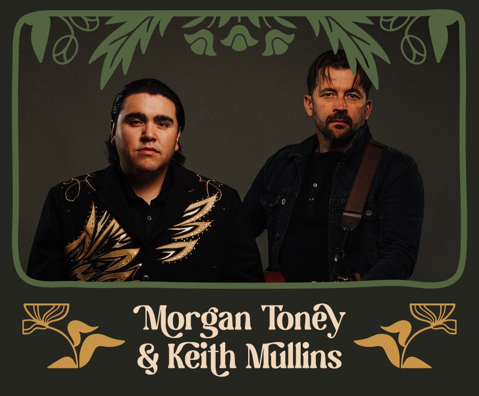 Morgan Toney sitting beside Keith Mullins. Morgan is a young man and has medium length dark hair that is slicked back. His shirt has images of golden feathers. Keith is a middle-aged man with short dark hair and a beard. He is holding an acoustic guitar.