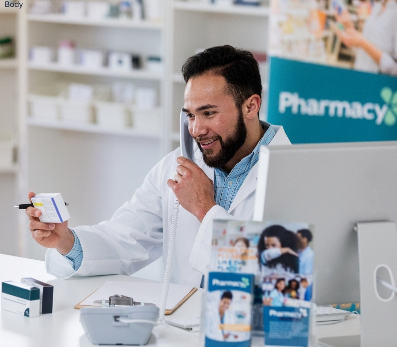 A male pharmacist with tan skin, dark hair, and a beard. He is looking at a package of medicine while talking on the phone.